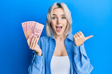 Sticker - Young blonde girl holding thai baht banknotes pointing thumb up to the side smiling happy with open mouth