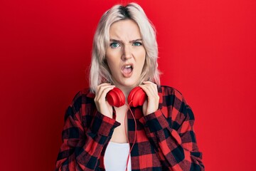 Sticker - Young blonde girl listening to music using headphones in shock face, looking skeptical and sarcastic, surprised with open mouth