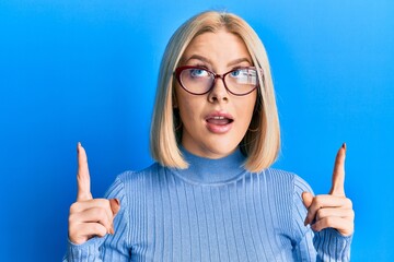 Young blonde woman wearing casual clothes and glasses amazed and surprised looking up and pointing with fingers and raised arms.