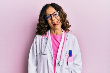 Middle age hispanic woman wearing doctor uniform and glasses sticking tongue out happy with funny expression. emotion concept.