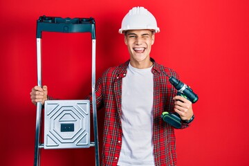 Wall Mural - Young hispanic man holding screwdriver wearing hardhat by construction stairs winking looking at the camera with sexy expression, cheerful and happy face.