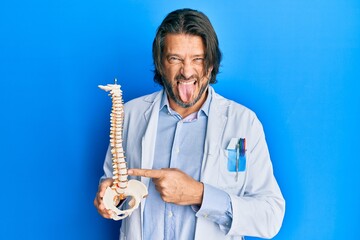 Sticker - Middle age handsome traumatologist man holding anatomical model of spinal column sticking tongue out happy with funny expression.