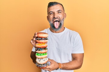 Middle age handsome man holding tasty colorful doughnuts sticking tongue out happy with funny expression.