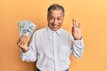 Canvas Print - Middle age indian man holding dollars doing ok sign with fingers, smiling friendly gesturing excellent symbol