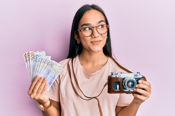 Sticker - Young asian woman holding vintage camera and swedish krona smiling looking to the side and staring away thinking.