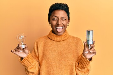 Wall Mural - Young african american girl holding lightbulb for inspiration and idea sticking tongue out happy with funny expression.