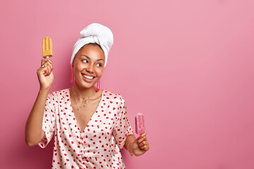 Wall Mural - Dark skinned cheerful housewife chills at home with tasty ice cream dances carefree has good mood dressed in silk pajama wears bath towel on head isolated over pink background empty space aside