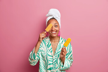 Wall Mural - Positive ethnic housewife with dark skin covers eye with tasty cold ice cream has fun while eating delicious sweet dessert wears domestic clothes wrapped bath towel on head. Summer time concept