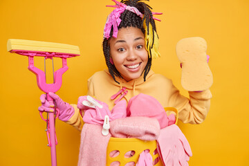 Wall Mural - People householding and spring cleaning concept. Smiling dark skinned woman poses with mop and sponge brings house in order does laundry wears protective rubber gloves isolated on yellow wall