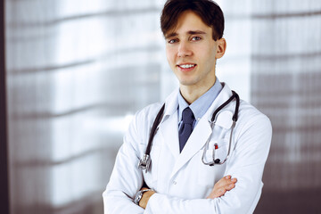 Cheerful smiling man-doctor standing with arms crossed in clinic. Perfect medical service with young smart physician in hospital. Medicine concept