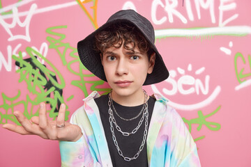 Poster - Cool looking indignant youngster shrugs shoulders and looks clueless at camera dressed in fashionable clothes poses against graffiti wall poses at urban place. Youth lifestyle and hooliganism