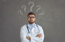 Smart Doctor In White Lab Coat And Glasses With Question Marks Above Head Standing Arms Folded And Thinking About Treatment Plan, Doubting Diagnosis, Looking For Solution To Patient's Health Problem