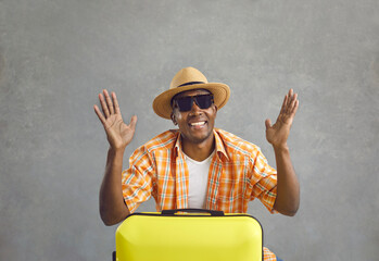 Wall Mural - Studio portrait of happy cheerful black tourist in sunglasses and sun hat with yellow travel suitcase. African American man going on summer getaway trip gesturing as he tells about his vacation plans