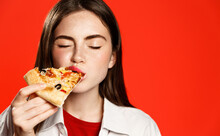 Photo Of Teenager Enjoys Delicious Slice Of Pizza, Likes This Taste, Closes Eyes From Pleasure, Has Good Appetite, Dressed In Casual White Shirt, Isolated Over Red Background. Hungry Woman Indoor