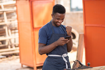 Wall Mural - excited african carpenter looking at his phone, seeing good news and being happy about it