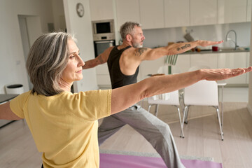 Wall Mural - Fit middle aged 50s family couple doing warrior yoga exercise at home. Sporty healthy old mature man and woman doing fitness workout standing in living room. Active seniors sport training concept.