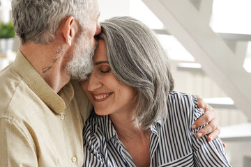 Wall Mural - Happy affectionate mature adult husband embracing kissing wife enjoying tender moment or love and care. Mid age 50s romantic grey haired married couple bonding together, hugging at home. Close up