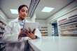 © StratfordProductions - Low angle view of young female woman wearing labcoat working in chemist using digital tablet to confirm online order