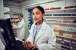 © StratfordProductions - Young woman working in pharmacy typing on computer while checking inventory wearing labcoat standing behind counter