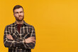 Thoughtful sceptical young man in checkered shirt with beard keeps arms crossed and thinking over yellow background Looking away to the side
