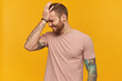 © timtimphoto - Disappointed embarrassed young bearded man with tattoo and eyes closed in pink tshirt keeps hand on forehead and looks upset over yellow background