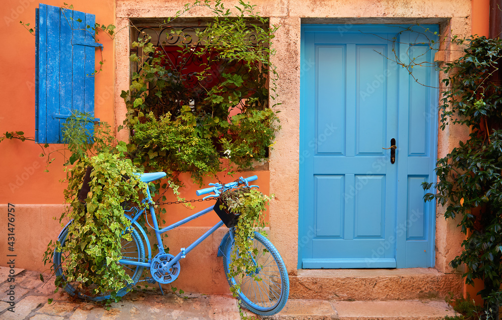 Rovinj, Istria, Croatia. Old blue bicycle at street near door of entrance in house among green bushes and flowerpots with flowers. Picturesque cosy lane. Window shutters on windows. - obrazy, fototapety, plakaty 