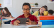 © TommyStockProject - Cheerful african-american boy in headphones holding tablet while sitting at desk in school classroom