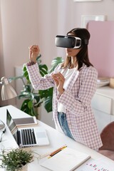 Canvas Print - Confident young woman in virtual reality headset pointing in the air while standing at her working place in office