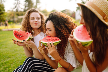 Three young having fun eating watermelon In the park. Young friends laughing and enjoying holidays together. Friendship, youth and travel concept.