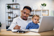© sofiko14 - Focused young man working on laptop and writing on clipboard while sitting at table with baby boy on knees. African father taking care of his son and working from home.