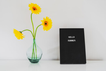 Quote - Hello summer on the Letter board and fresh yellow gerbera flowers in the glass vase on a white table. Sunny summer mood background. Minimalism. Copy space