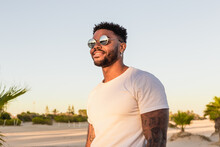 Portrait Of A Handsome Black And Young American Man Wearing Sunglasses And Smiling During Sunset At The Beach
