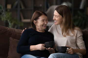 Wall Mural - Smiling grownup Caucasian female kid child relax rest on couch with mature 60s mom drink coffee tea together. Happy two generations of women enjoy family reunion at home. Relation concept.