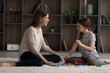 © fizkes - Little teen girl child and young Caucasian mother or nanny sit on floor at home play on weekend together. Happy mom and small teenage daughter have fun engaged in funny game activity in living room.