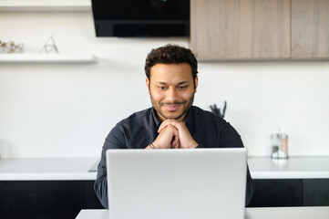 Front view of positive hindoo man in smart casual shirt using laptop sitting at the desk in the kitchen, young indian male student watching webinars, educational courses, learning on the distance