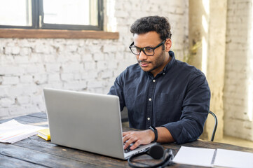 Focused hindu freelancer guy wearing eyeglasses using laptop in contemporary office space, concentrated mixed-race man typing, messaging, indian programmer develops software, looks at computer screen