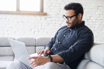 Serious multiracial man using laptop computer for remote work from home, hindu freelancer guy reading incoming emails, chatting online, researching or studying online sitting on the couch at home