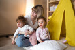 © Tatsiana - a happy mother with two twin girls plays at home on the floor sitting near a yellow teepee.