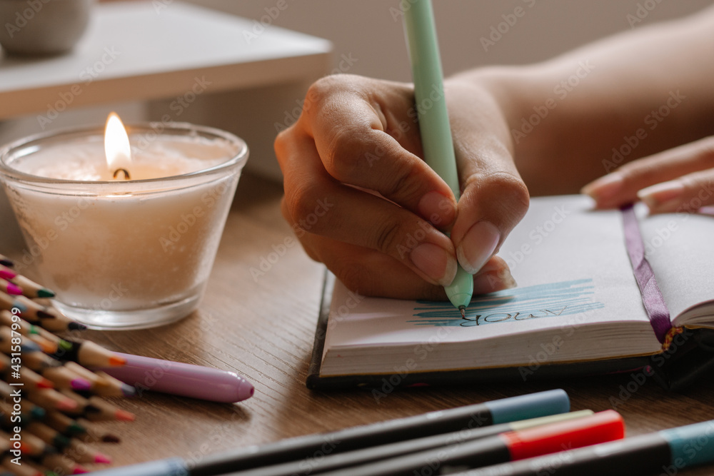 mano de mujer bocetando sobre cuaderno blanco ,  usando marcadores y lapiceros , decorado con colores cálidos madera y vela encendida. - obrazy, fototapety, plakaty 