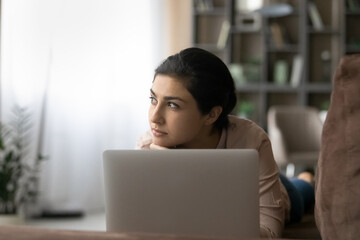 Close up thoughtful Indian woman lying on couch with laptop at home, distracted from device, looking to aside, pensive young female freelancer or student pondering project strategy, planning
