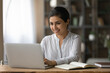 © fizkes - Smiling Indian woman using laptop, studying online at home, sitting at desk with books and computer, working on research project or homework, positive businesswoman writing report or email