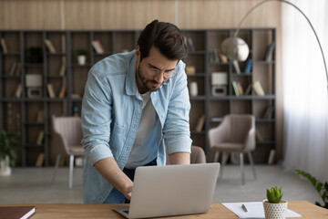 Wall Mural - Confident businessman in glasses working on laptop, typing, standing at home office desk, serious focused young man looking at screen, freelancer reading or writing business email, research project