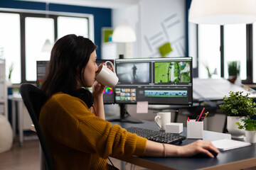 Wall Mural - Professional colorist working in video footage using post production software, drinking coffee. Videographer editing audio film montage at pc with two monitors, sitting on desk in creative agency