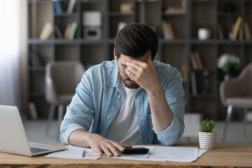 Unhappy businessman in glasses calculating bills, worried by financial problem, unexpected debt, bankruptcy, frustrated young man sitting at desk with laptop and calculator, lack of money concept