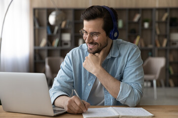 Close up smiling man in headphones and glasses using laptop, writing taking notes, motivated student watching webinar or training, listening to lecture, learning language, studying online at home
