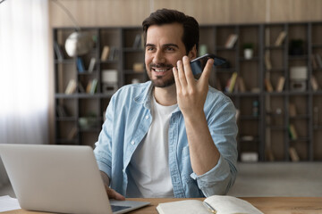 Wall Mural - Smiling businessman recording audio voice message on smartphone, speaking, sitting at work desk with laptop, positive young man chatting online by speakerphone, activating digital assistant on gadget