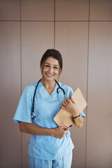 Wall Mural - Cheerful female doctor with clipboard standing in clinic