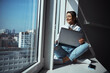 © Svitlana - Joyful woman sitting on windowsill and using laptop
