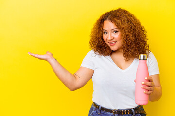 Wall Mural - Young latin woman holding a thermos isolated on yellow background showing a copy space on a palm and holding another hand on waist.