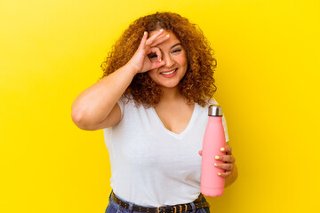 Wall Mural - Young latin woman holding a thermos isolated on yellow background excited keeping ok gesture on eye.